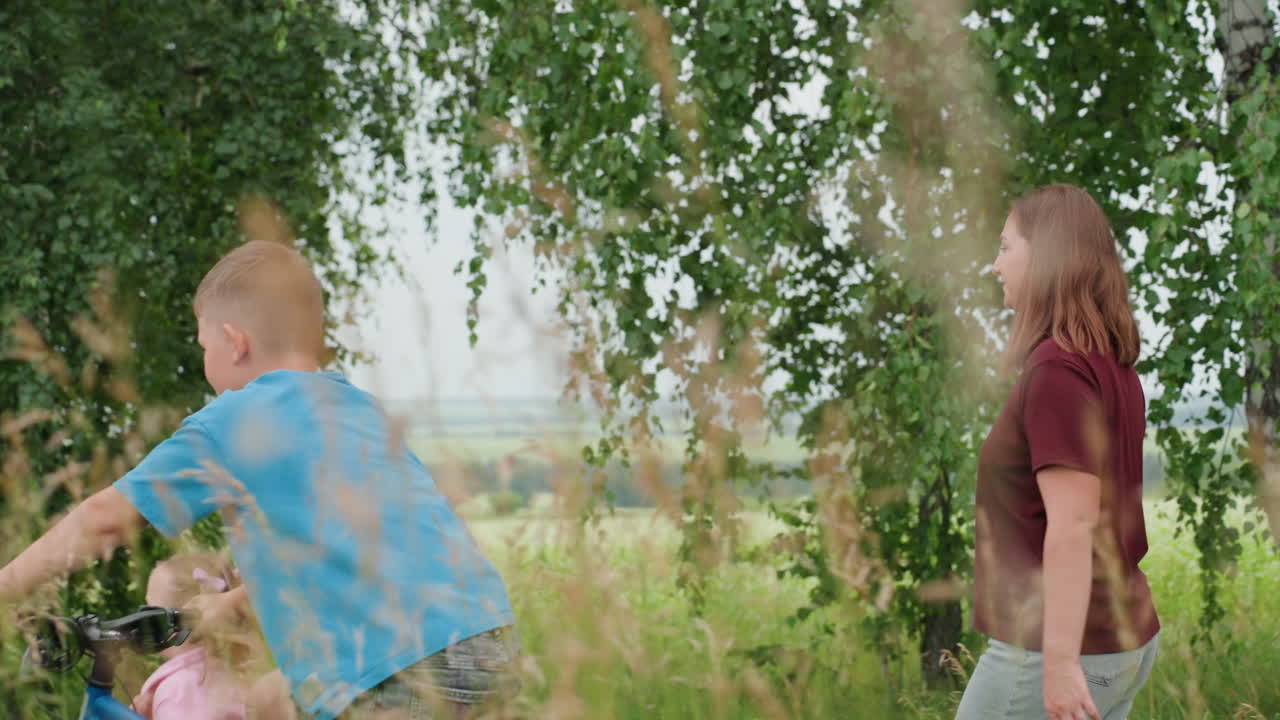 하얀 피부의 엄마와 아이들이 초원 길을 걷고 있어요. 엄마는 어린 딸을 이끌고, 큰 아들은 페달을 밟으며 지나가고, 십 대 아이는 뒤따르고 있네요. 자작나무와 키 큰 풀이 아름다운 시골 풍경을 감싸고 있으며, 부드러운 햇살이 비추고 있어요.