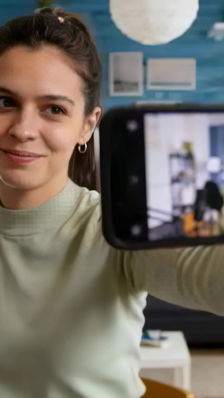 Video verticale: Donna che tiene il telefono, inizia un selfie a casa, aggiustando la posa, con un maglione verde chiaro.
