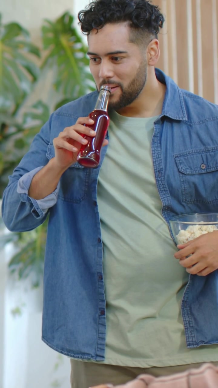 Video verticale: Uomo che ride in camicia di jeans sorseggia da una bottiglia a casa, tiene popcorn e reagisce a un amico.