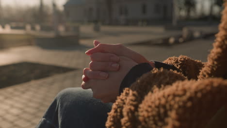 primer plano de una persona sentada con la cara no visible, con pantalones vaqueros azules y un abrigo marrón. las manos están fuertemente juntas, lo que sugiere un pensamiento profundo y meditación