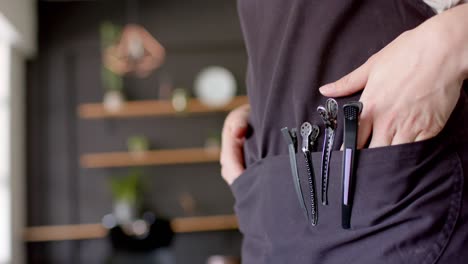 Midsection-of-caucasian-female-hairdresser-in-apron-with-hands-and-hair-clips-in-pocket,-slow-motion