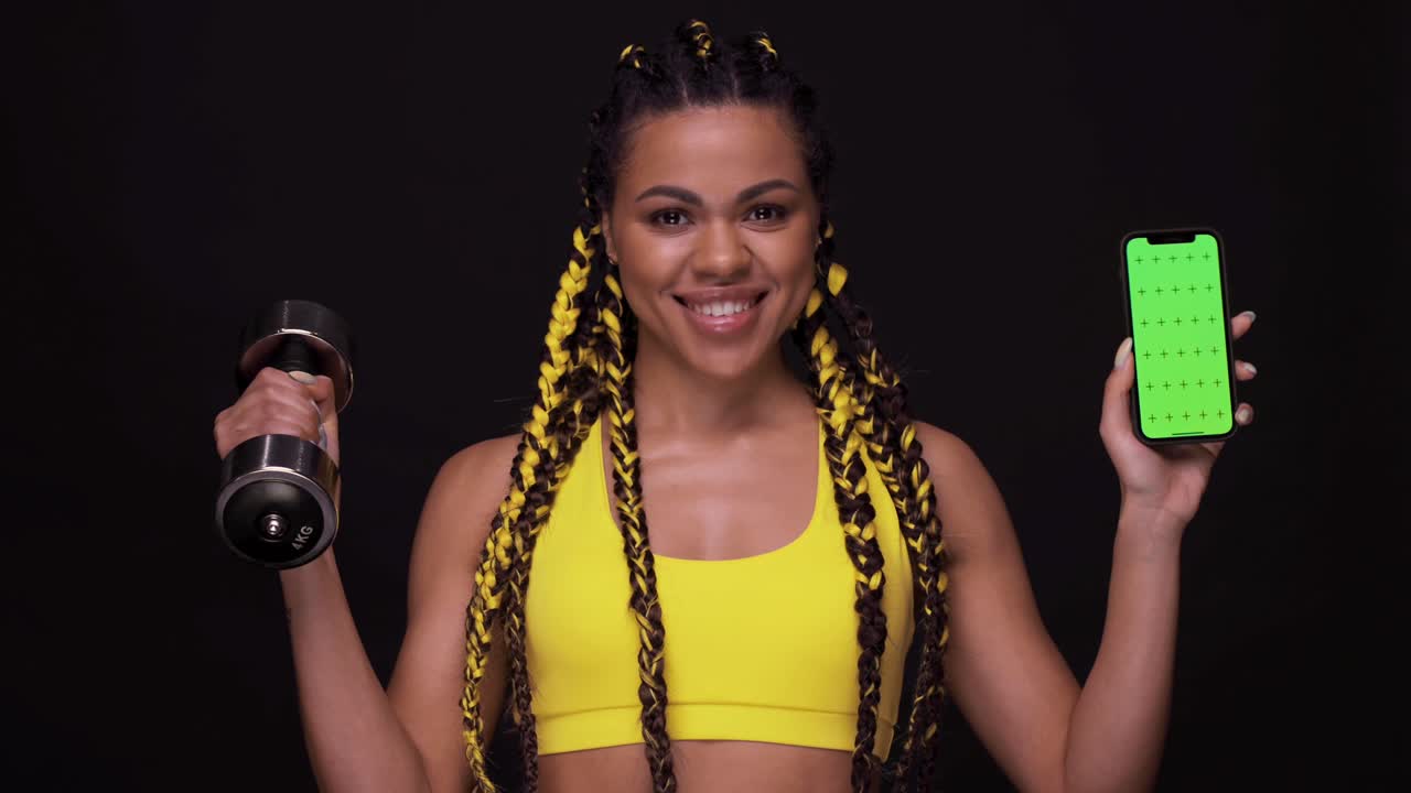 Portrait of a smiling afro american girl choosing between mobile phone and dumbbell. The screen with chroma key. Woman isolated over black background