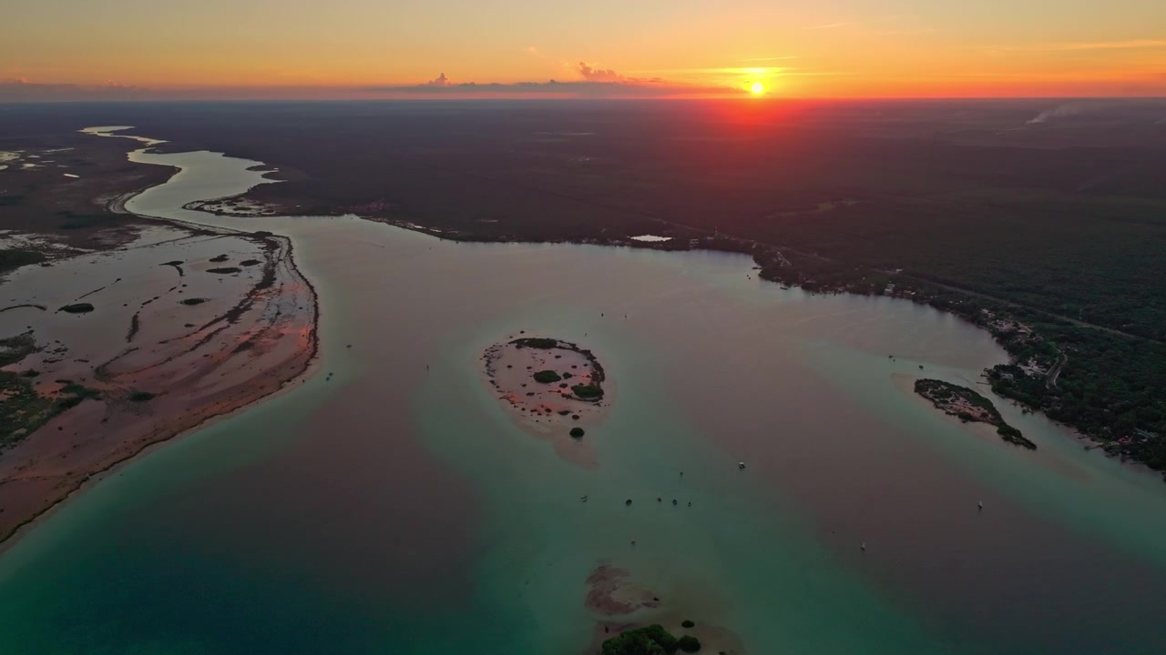 Vista aerea al tramonto su un canale tortuoso della laguna di Bacalar, Messico, con il sole arancione all'orizzonte riflesso nell'acqua calma e immobile
