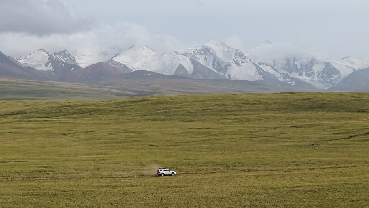 Fordon som kör genom en vidsträckt, öppen äng med snötäckta berg i bakgrunden i Kirgizistan. – flygbild