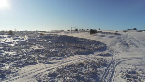 Snow-covered-rural-winter-countryside-track-footprint-shadows-terrain-aerial-view-panning-right-shot