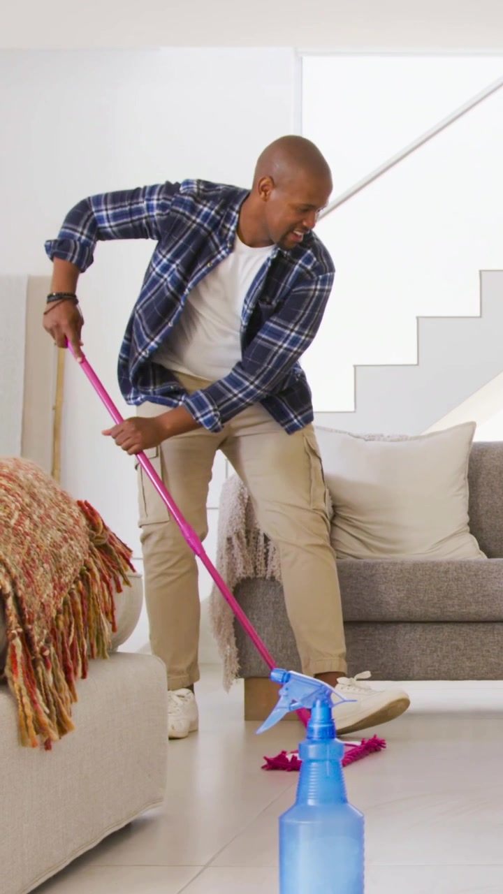 Vertical video: Starting holding pink mop, man in plaid cleaning living room floor with blue bottle