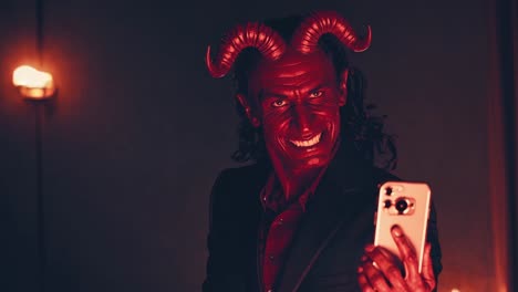 red devil businessman with horns is taking a selfie with smartphone in elegant suit in a dark room illuminated by a torch, smiling ironically
