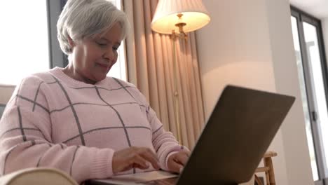 mujer biracial senior feliz sentada en un sillón y usando una computadora portátil, inalterada, en cámara lenta
