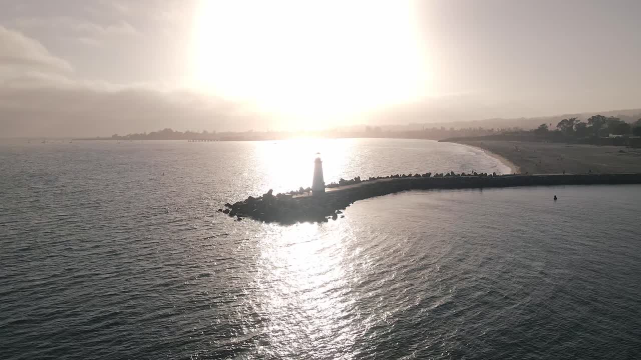 A slow motion drone shot circling the Walton Lighthouse and the small crafts harbor in Santa Cruz California with the sun setting in the background with sailboats in the Pacific Ocean