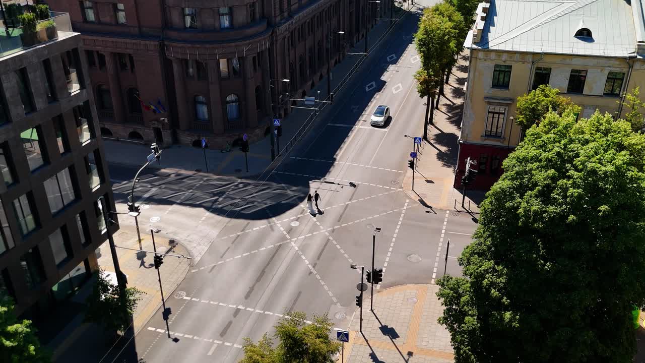 Aerial view of a quiet Kaunas city intersection with minimal traffic. Pedestrians cross the street, surrounded by trees and historic buildings under clear, sunny weather. Urban life in a calm moment.