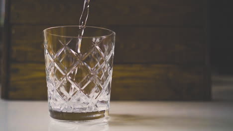 Slow-motion-close-up-shot-of-tonic-water-being-poured-into-a-fancy-glass-against-a-wooden-background-with-a-reflection-on-the-white-table,-alcoholic-drink