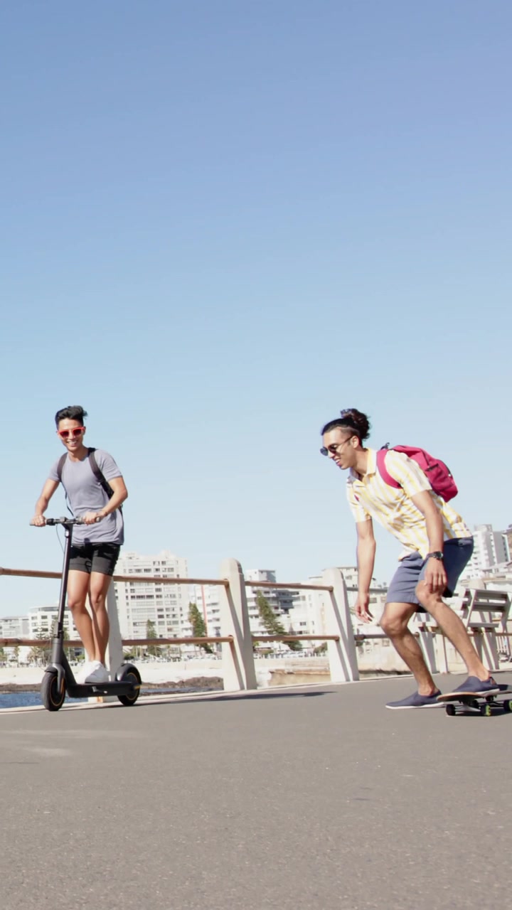 Video verticale: Amici che si avvicinano da sinistra in scooter e spingendo un longboard per svago.