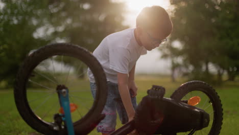 a young boy is seen intently rotating the pedals of an upside-down bicycle, causing the tire to spin in a grassy field