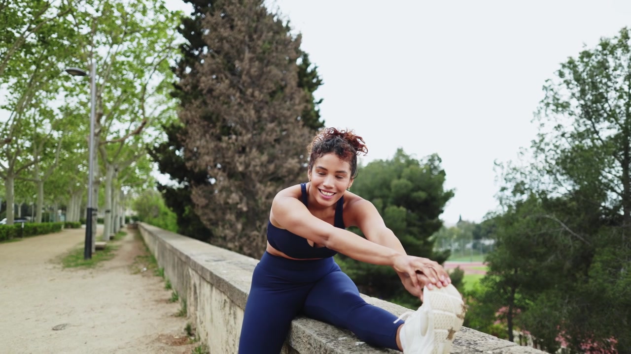 Young Woman in Sportswear Stretching Leg on Wall in Park