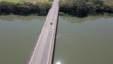 Old-beetle-green-car-traveling-passing-by-the-bridge-over-the-river-aerial-view-in-a-sunny-day-with-sun-reflexion