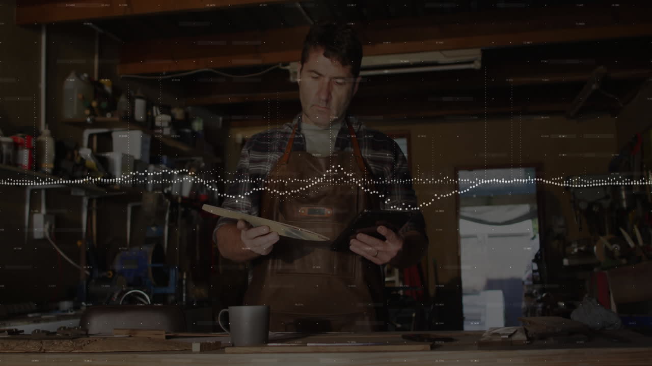 Mature craftsman standing behind workbench, examining plank and viewing tablet with business graphs