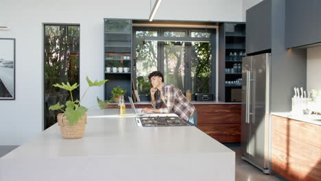 using laptop in modern kitchen, teenage boy leaning on counter and thinking