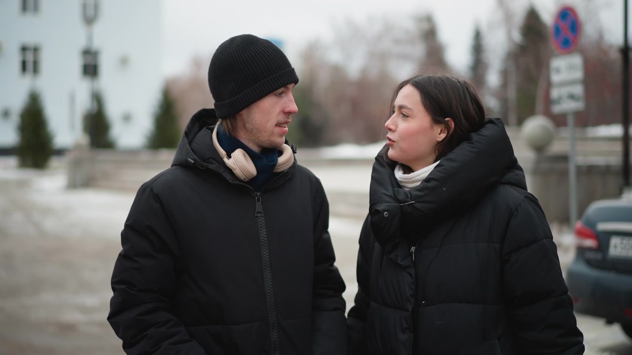 Close up of man and woman walking outdoors in winter holding hands. Both wear black puffer jackets man has beanie with headphones around neck while woman wears white scarf. Background shows