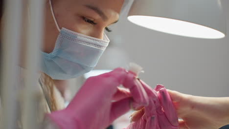 closeup view of nail technician wearing face mask and pink gloves carefully grooming customer's nails under bright white lamp, with strong focus on precision, hygiene, and personal care details