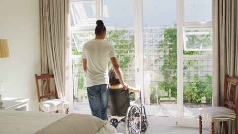 mujer biracial en silla de ruedas y pareja masculina mirando por la ventana en un dormitorio soleado