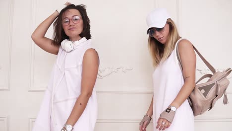 two female hipsters in white dresses dancing on the background of white wall. front footage. slow motion