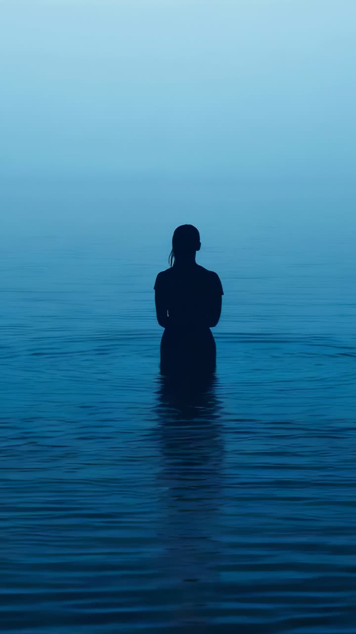 Vertical video: Haze drawing woman in shirt standing in calm water, showing reflection and ripples