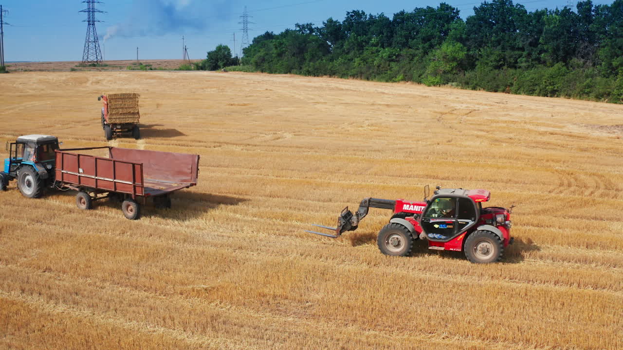 Skid loader follows the empty tractor machine in the field. Loader goes to a hay bale to pick it up. Blue sky backdrop.