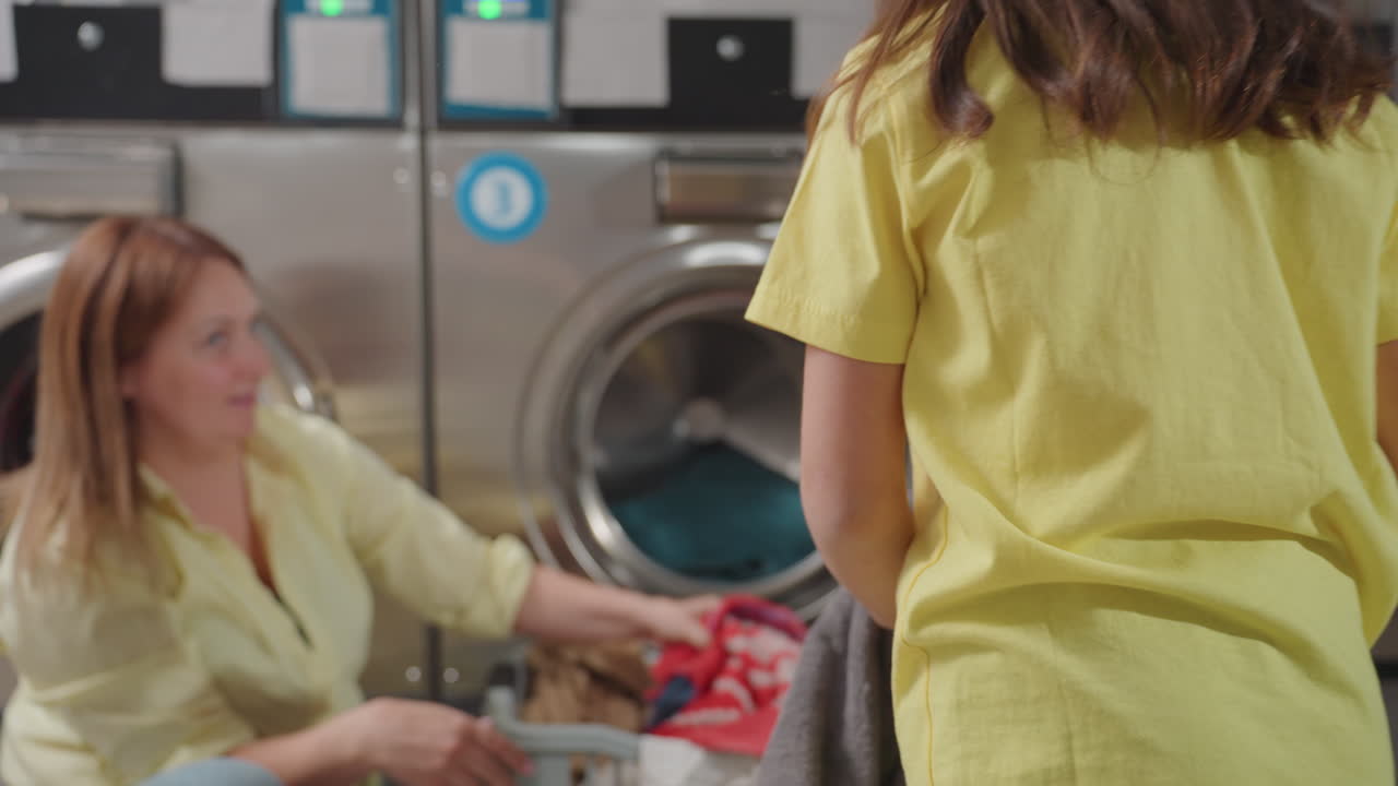 Mother with toddler packing clothes into washer at laundromat, woman kneels by open stainless machine, child gestures while basket of towels waits, sorting textiles, clean service routine inside