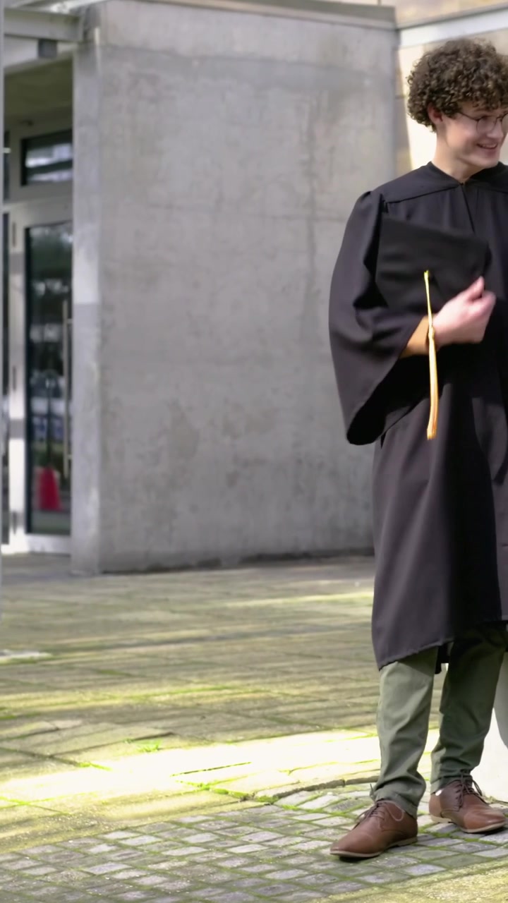 Video verticale: Un laureato/a in toga nera chiacchiera nel campus dopo la cerimonia, tenendo in mano il tocco e un caffè.