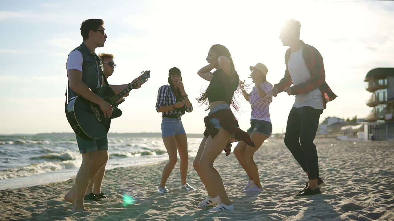 Summer party on the beach. Young friends drinking cocktails, dancing in the cirkle, playing guitar, singing songs and clapping on a beach at the water's edge during the sunset. Slowmotion shot