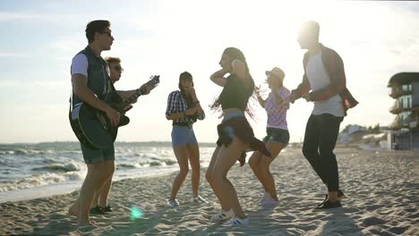 summer party on the beach. young friends drinking cocktails, dancing in the cirkle, playing guitar, singing songs and clapping on a beach at the water's edge during the sunset. slowmotion shot