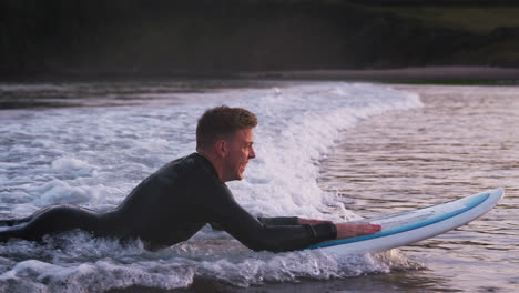 man wearing wetsuit lying on surfboard and riding wave into shore