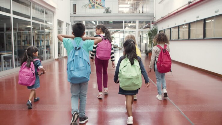Vista trasera de un grupo feliz y multiétnico de escolares con mochilas coloridas caminando por el pasillo de la escuela.