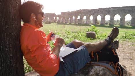 ciclista con bicicleta sentado en una roca bajo un árbol escuchando música con su portátil y bebiendo agua frente al antiguo acueducto romano. joven atractivo hombre atlético con auriculares deportivos naranjas y mochila en el parco degli acquedotti en roma