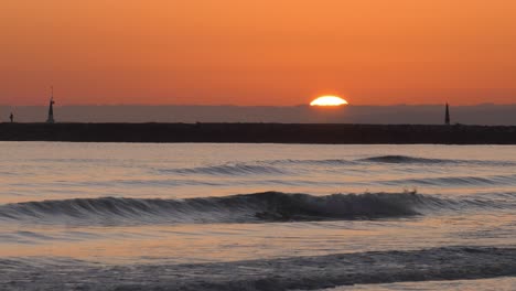 Sun-rising-above-cloud-into-orange-sky-over-gentle-waves-at-dawn,-man-silhouetted-on-sea-wall,-mediterranean,-spain