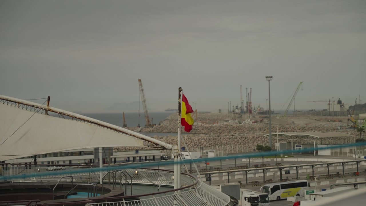 Bandiera spagnola sventola su un terminal portuale industriale con gru edili e frangiflutti in Spagna.