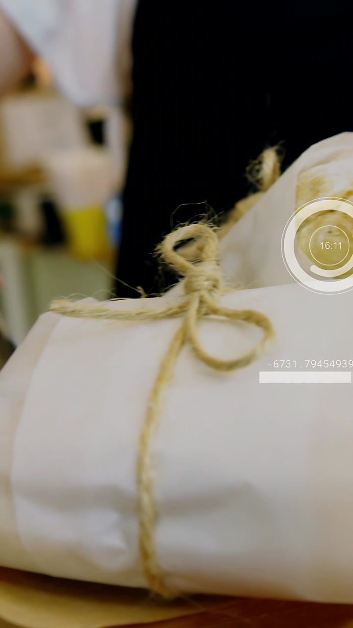 Vertical video: Hand with tongs nearing and adjusting twine-wrapped sandwiches for display with HUD