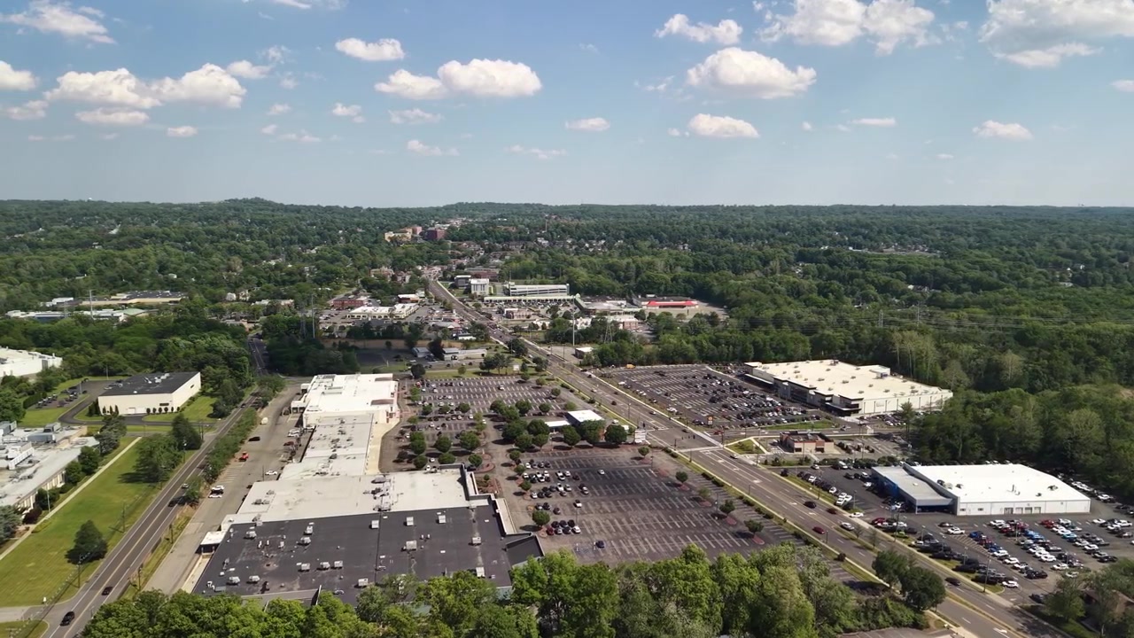 Aerial perspective showing bustling suburban shopping centers, expansive parking lots, and flowing traffic on a major highway, all set against a backdrop of lush green trees on a sunny day