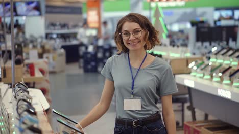 Positives-Weibliches-Verkäufer--Oder-Verkäuferporträt-Im-Supermarktgeschäft.-Frau-In-Blauem-Hemd-Und-Leerem-Abzeichen,-Die-In-Die-Kamera-Schaut-Und-Lächelt.-Elektronische-Geräte-Im-Hintergrund.-Zeitlupe