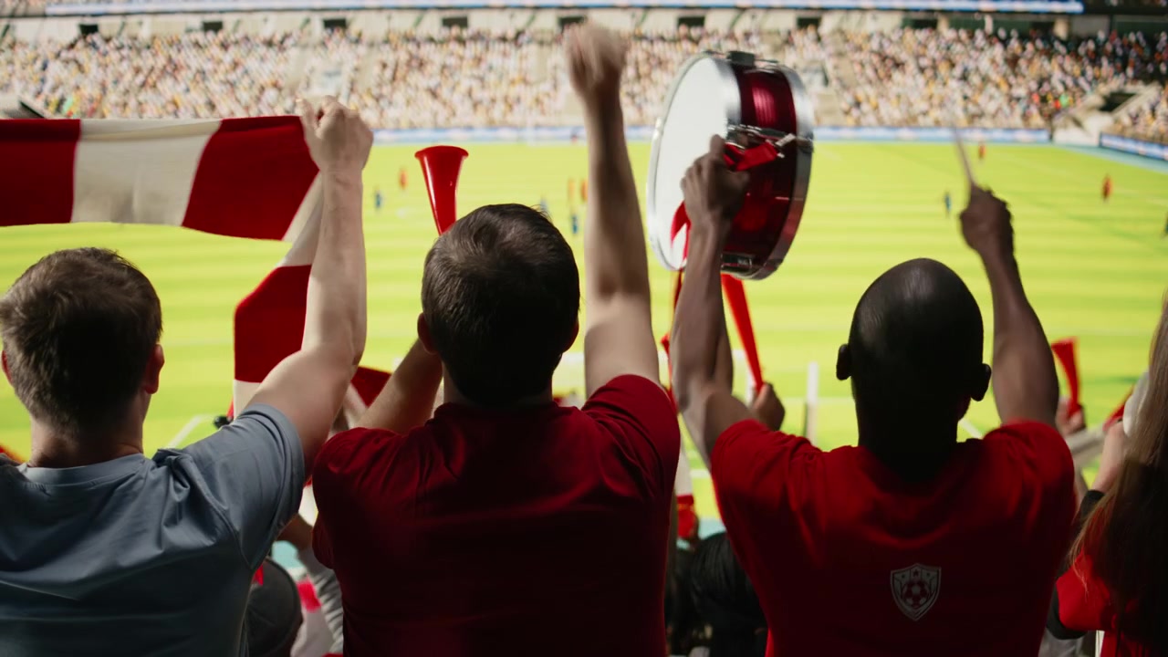 Vista trasera de aficionados al fútbol con camisetas rojas observando el campo desde las gradas
