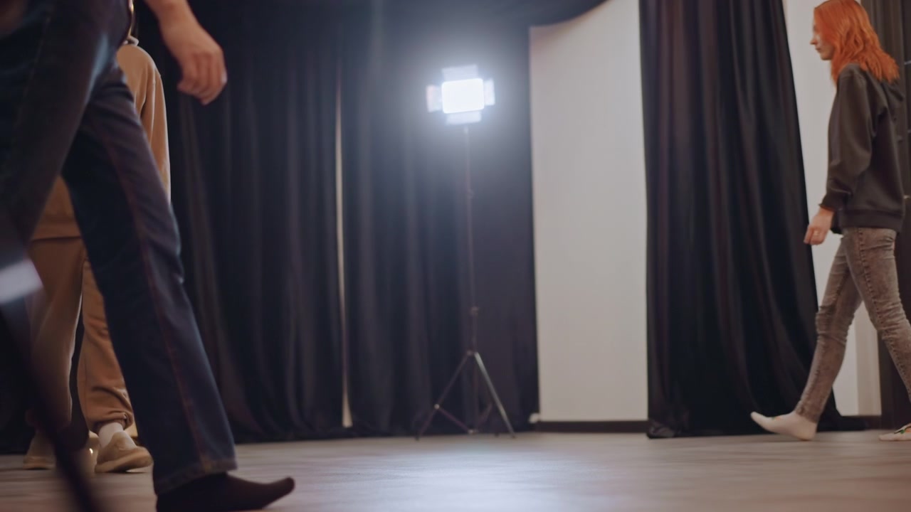 Studio rehearsal with white students walking across stage under soft lights, acting coach directing blocking and footwork, casual clothing and sneakers, black curtains and spotlight, collaborative