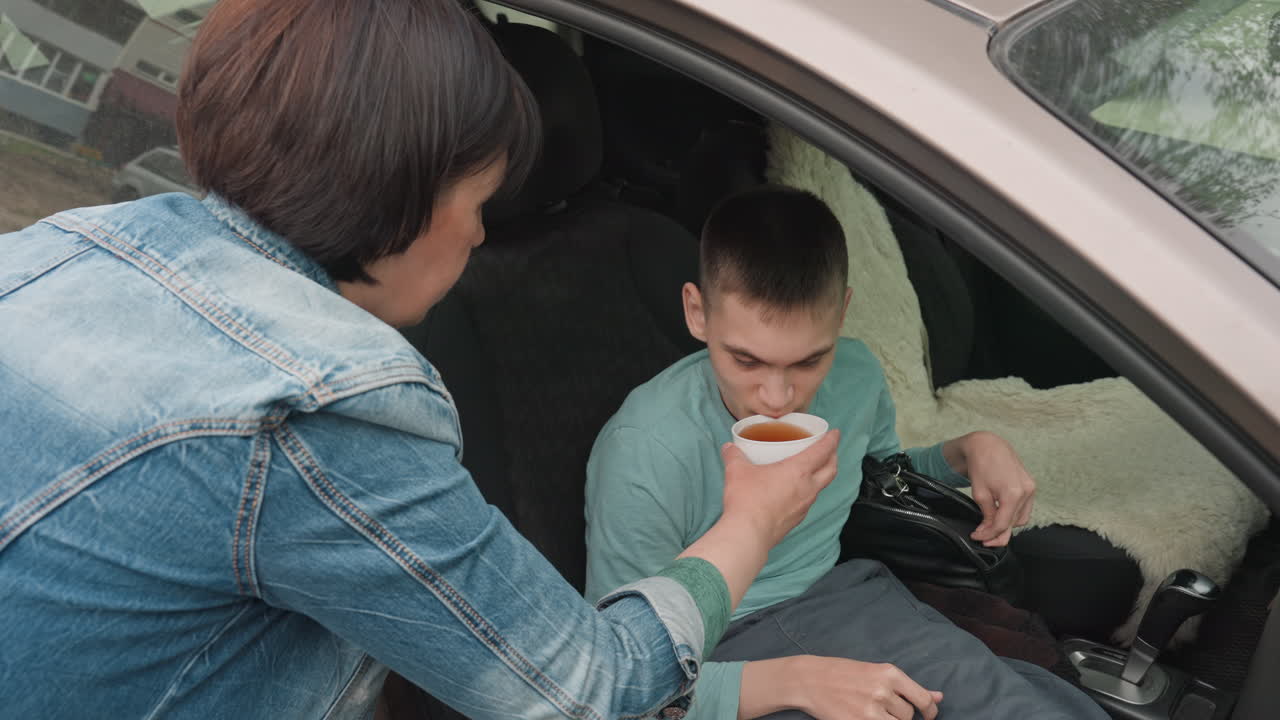 Teen Sipping From Cup In Parked Car As Mother Caregiver Offers Drink, Careful Hand Positioning And Supportive Assistance, Denim Jacket, Seatbelt And Wheelchair Visible, Calm Daytime Scene Emphasizing
