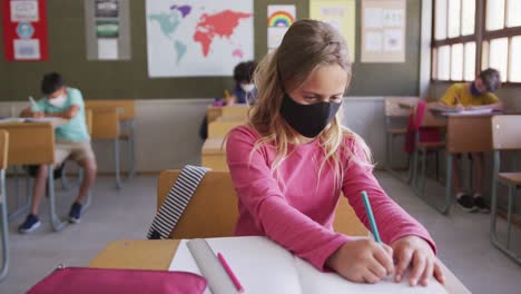 Mädchen-Mit-Gesichtsmaske-Schreibt,-Während-Sie-In-Der-Schule-Auf-Ihrem-Schreibtisch-Sitzt