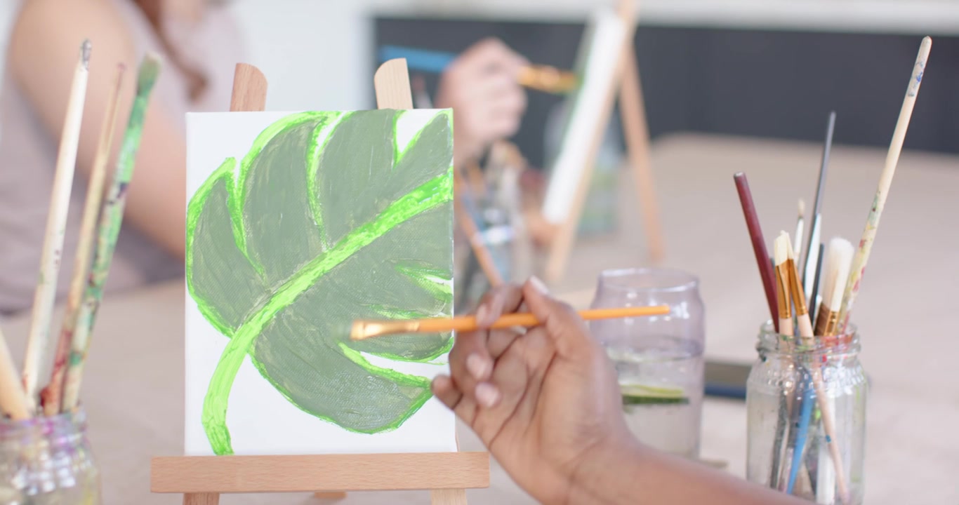 Mujeres afroamericanas pintando en un estudio, una mano sumergiendo el pincel para terminar una hoja de neón en un lienzo.