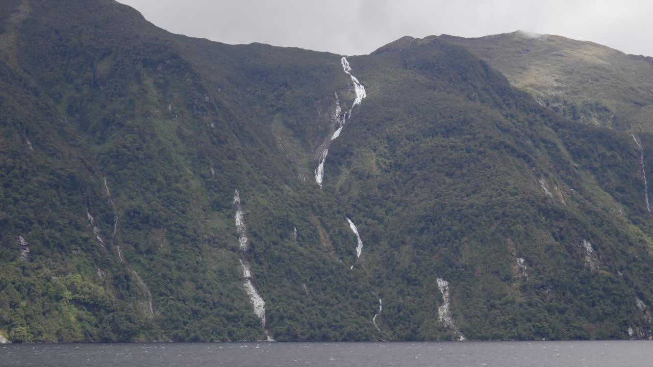 Una lunga cascata scende lungo una ripida montagna a Doubtful Sound, scavando attraverso roccia e nebbia in una potente dimostrazione della bellezza della natura selvaggia e remota