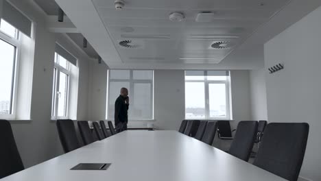 a man paces and converses on his mobile phone in front of an empty conference room table, surrounded by chairs, reflecting the concept of business communication and anticipation