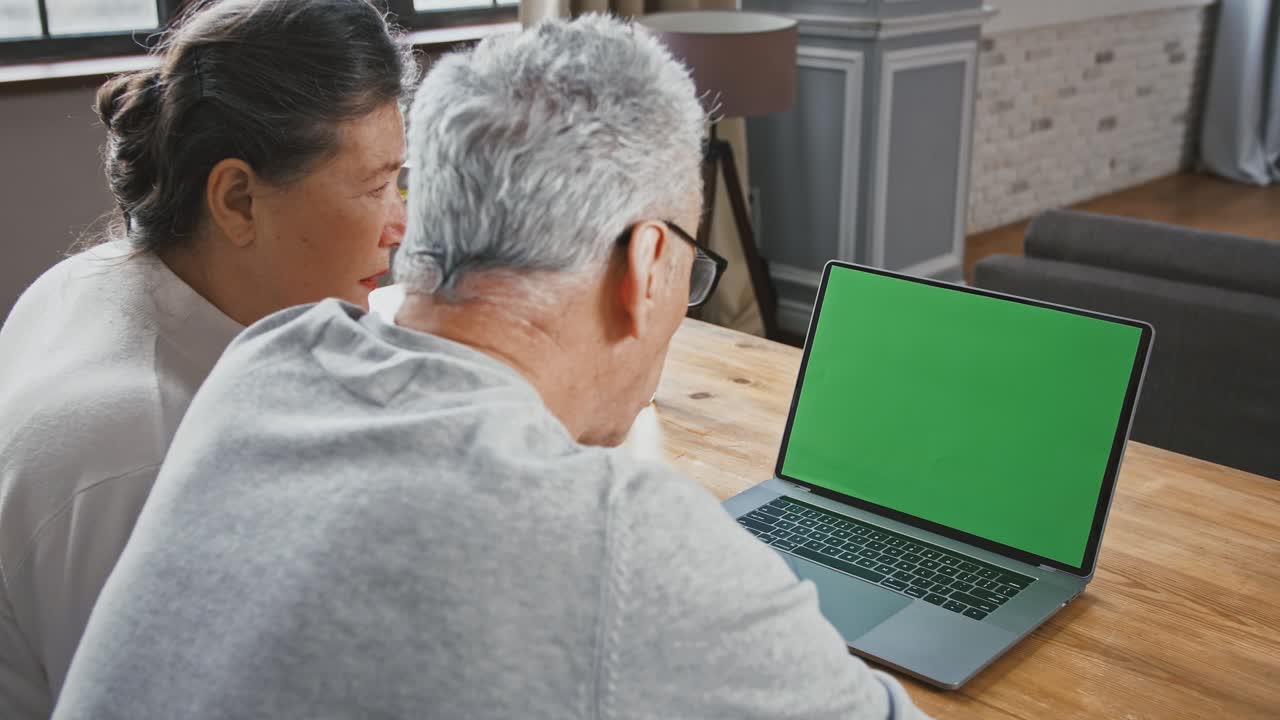 vista trasera de una pareja de ancianos mirando la pantalla verde de una computadora portátil y hablando con alguien usando una llamada de video en línea, clave de croma