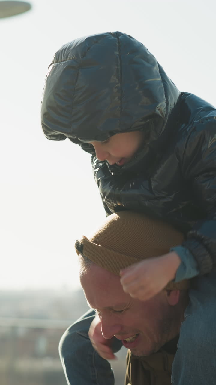 un primer plano de un padre llevando a su hijo en sus hombros, mientras el niño juega con la capucha de su padre, el padre lleva una chaqueta marrón, y el hijo lleva una chaquette negra, con una vista borrosa del paisaje urbano