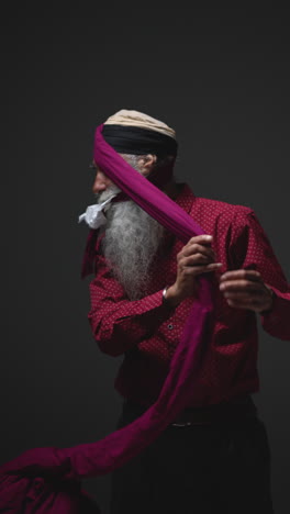 vertical video low key studio lighting shot of senior sikh man folding fabric for turban against plain dark background 2