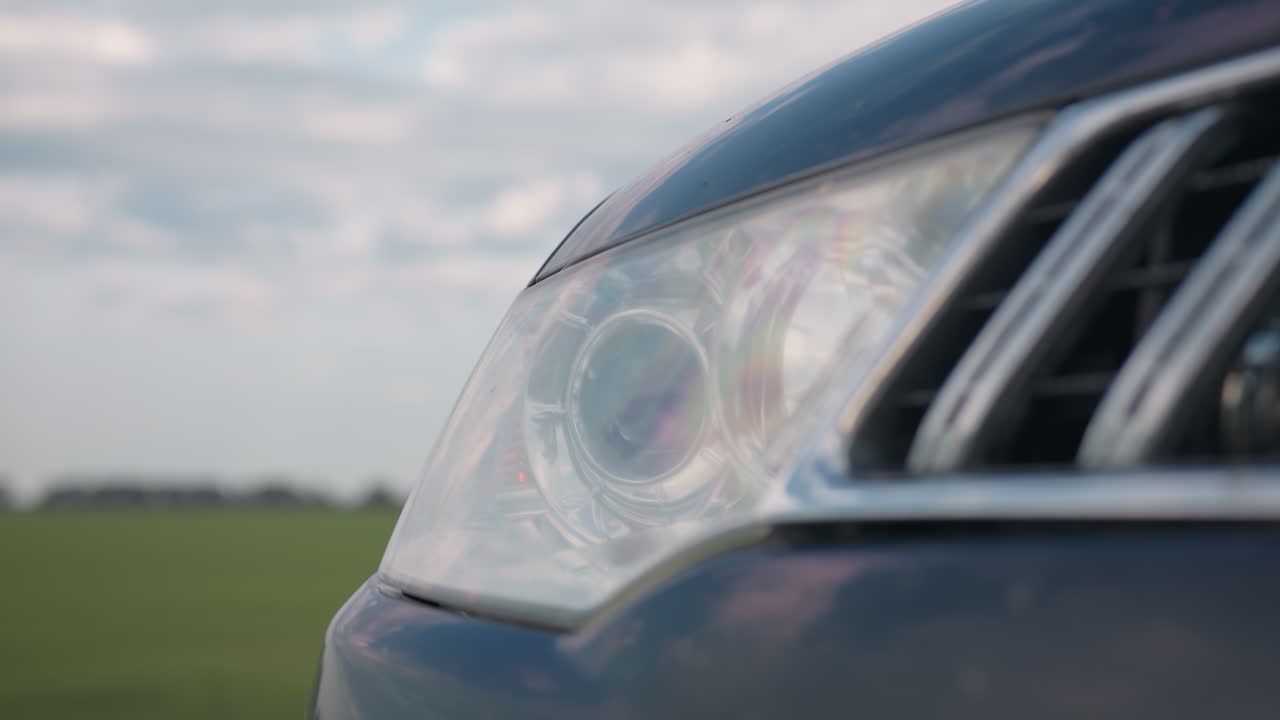 Close up of modern car headlight blinking gently, with chrome grille catching light reflections, positioned beside open green field under cloudy sky, rural setting softly blurred in background
